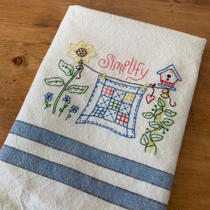 Embroidered tea towel that shows a quilt, a bird house, sunflower, and the word Simplify