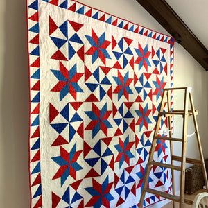 Red, White, and Blue Bombs Burst quilt hanging on a wall with a ladder in front of it.