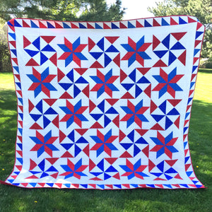 Bombs Bursting Quilt, in red, white and blue, hanging in a park