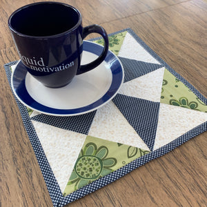 Pin Wheel Mug Rug Mini Quilt in White, Blue and Green, sitting on the table with a cup and plate